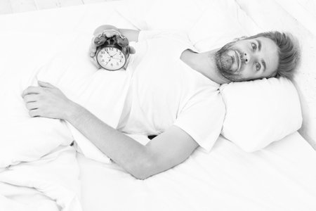 Morning awakening. Sleepy man turning off alarm clock. Waking up early. Time to wakeup and get up. Morning wakeup. Sleepy man with alarm clock in hand while lying in bed. Sleep disorderの写真素材