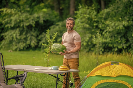Camp and nature picnic. Man camping. Camp watermelon picnic. Yummy healthy food. Fresh food in camp. Man holding watermelon in summer camping. Man hiker on summer day. Eat fruit outdoorの写真素材