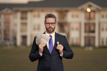 House owner hold money outdoor. Realtor offering new home. Broker make offer. Businessman broker with money. Successful realtor in suit with money dollar and house key. Commercial real estate brokerの写真素材