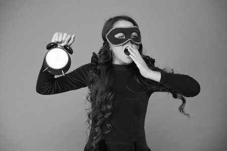 Girl late home after party celebration. End of holiday fun. Countdown. Girl being late and sleepy isolated on orange. Late time. Party countdown. Sleepy teen girl yawning in party mask with clockの写真素材