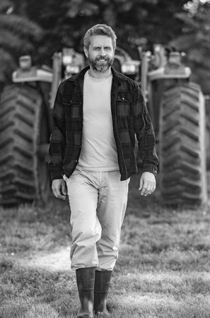 Farmer with harvesting machinery. Mature man at tractor. Tractor driver. Agriculture and field work. Farmer in field wearing gum boots with tractor outdoor. Crop harvesting. Agriculture machineの写真素材
