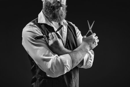 Handsome barber using retro blade and scissors while cutting hair. Modern barbershop haircut. Bearded hipster barber isolated on black. Man beard haircut by hairdresser at barbershop. Fresh haircutの写真素材