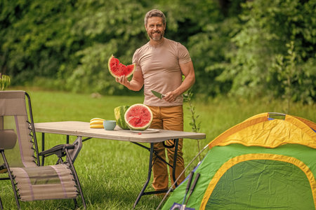 Man camping. Camp watermelon picnic. Yummy healthy food. Fresh food in camp. Man with watermelon in summer camping. Man hiker outdoor on summer day. Camp and nature picnic. Organic foodの写真素材