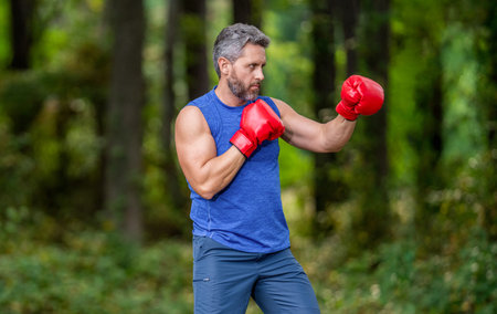 Strong athlete man. Sportsman in forest. Strong boxer in sportswear. Exercising and punching. Practicing boxing, relentless. Sportsman in boxing gloves outdoor. Mature man boxer. Sport man boxingの写真素材