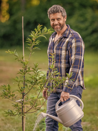 Gardener man gardening. Gardener in spring. Man with watering can in garden. Watering peach tree. Planting a tree. Gardening and watering orchard. Seedling for planting tree. Spring planting seasonの写真素材