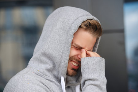 Young Caucasian man in hoodie suffering from headache or migraine pain outdoor