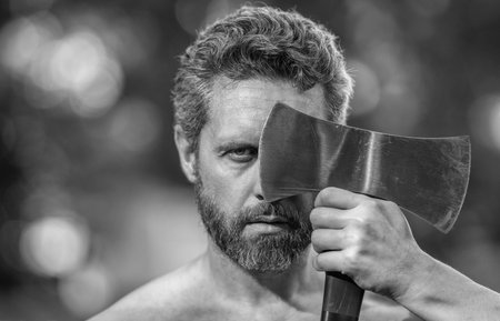 Lumberjack man face with axe. Lumberjack axe. Man with logging axe. Masculine man portrait. Lumberjack outdoor portrait.の写真素材