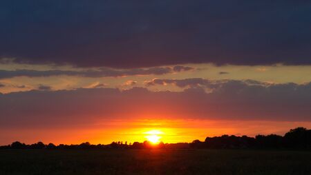 Langsam geht die Sonne unter am Horizont unter lila Wolken vor Silhuette von Wald und Feldの写真素材