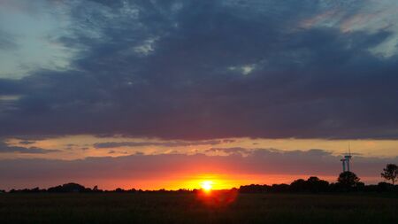 Langsam geht die Sonne unter am Horizont unter lila Wolken vor Silhuette von Wald und Feldの写真素材