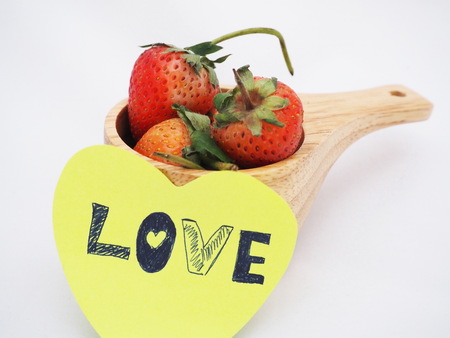 Love word was written on yellow paper and attached on wooden cup which fully contained strawberries on white background. Focusing on one strawberry in cup.の写真素材