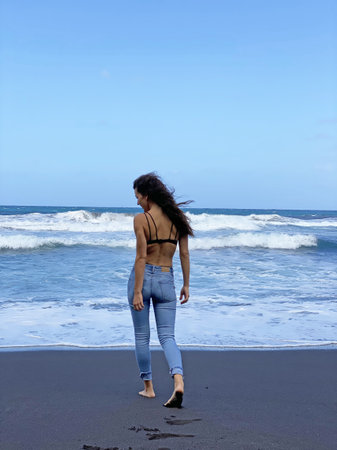 Young woman with black hair and blue jeans relaxing on the volcanic beach of the island of Lanzarote