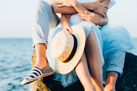 Close up of couple sitting on the stone near the sea or ocean. Photo of hands hugging and holding straw hat. Summer love concept. Holiday relaxing, beach vacation. Couple enjoying their time togetherの写真素材