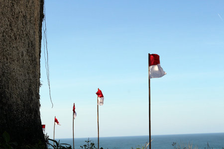 The Indonesian flag flies on a cliff with a backdrop of the beach and clear sky