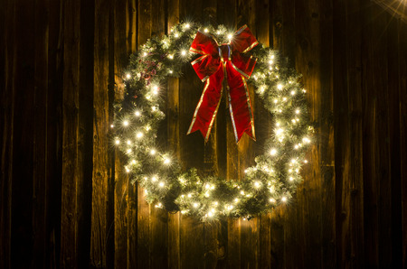 An old ranch barn is the perfect backdrop for a large lighted Christmas wreath, complete with red ribbon.  Photographed on a cold winter night, this wreath stands out against the weathered wooden background with plenty of copy space available for your ownの写真素材