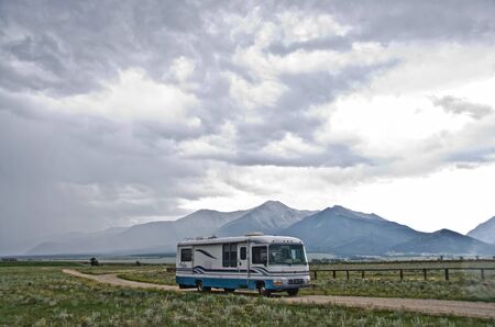 A large Class A motorhome drives down a dirt road at the base of Mount Princeton near Buena Vista, Colorado.  A summer rain storm is blowing in.のeditorial素材