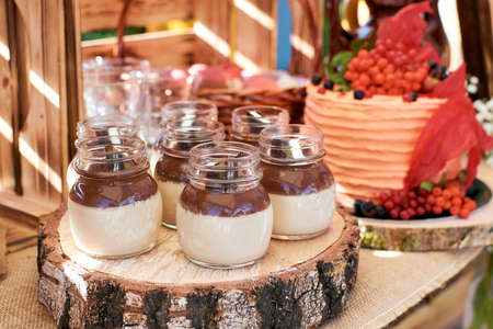 Chocolate cream dessert in a glass jar. Desserts stand on a wooden block, with a birthday cake in the background. Fragment of a sweet table in a rustic styleの写真素材