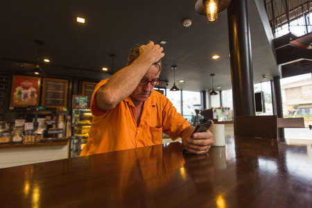 Man holding a mobile phone with his face in a cafe wearing a orange shirtの写真素材