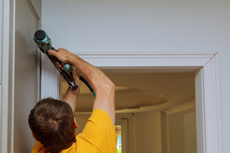 Carpenter brad using nail gun to moldings on doors, framing trim, with the warning label that all...