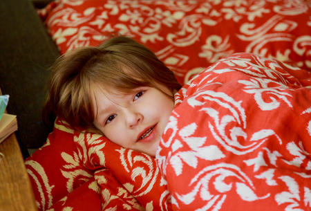 A small diseased girl in the bedroom. Little girl sitting on a bed wearing a pajamas Sick little girl lying in the bed with thermometerの写真素材