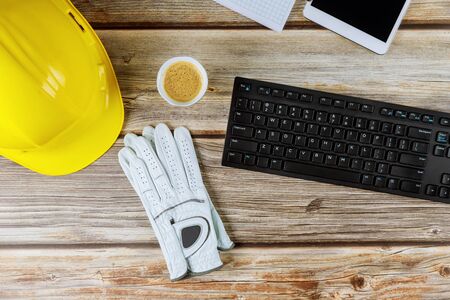 Yellow plastic safety helmet on construction builder office and leather work gloves computer keyboard wood backgroundの写真素材