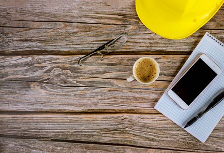 Workspace builders architect office table desk, blank open notebook with pen on yellow hard hat with cup of coffee, smartphone eyeglasses on wooden deskの写真素材