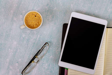 Working at office desk using a digital tablet with porcelain cup of black coffee, eyeglasses, weekly...