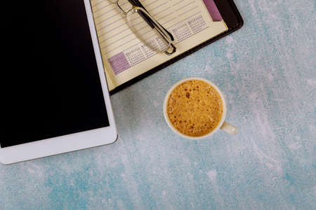 An open weekly notebook and digital tablet eyeglasses on the table a white cup of coffee.
