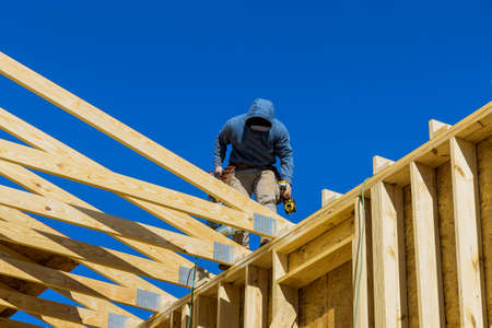 Builder on the unfinished roof with nailing wooden beams in frame house under construction with...