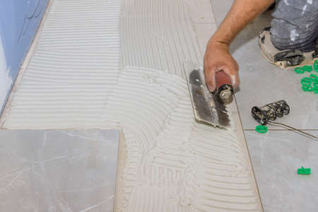 Reconstruction bathroom with worker using trowels to apply adhesive to concrete floors before tile...