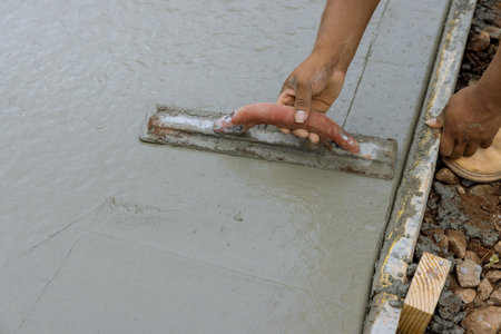 A construction worker is working on a new sidewalk that has just been poured on wet concrete while holding steel trowelの写真素材