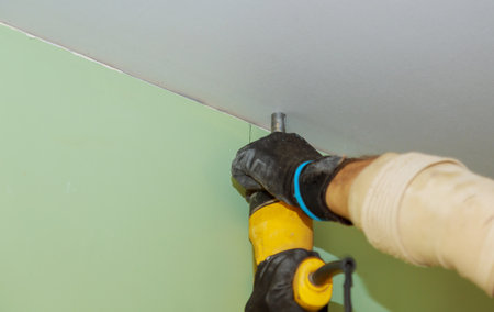 Using a screwdriver and drywall screws, worker screws the drywall into place on ceilingの写真素材