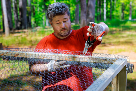 On farm worker is using scissors to cut metal grid wooden domestic chicken coops
