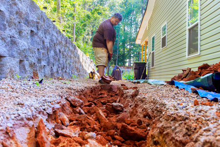 At construction site, worker digs trench for underground installation of drainage system using perforated pipes.の写真素材