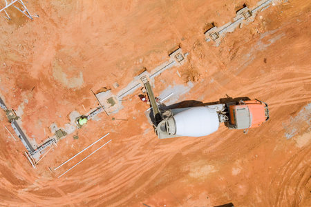Aerial view of construction site where concrete mixer truck is pouring fresh concrete into forms along foundation line on reddish soil.の写真素材