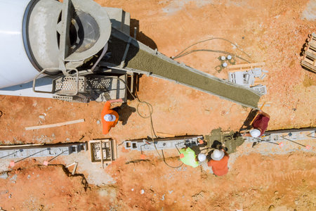 Construction worker pours concrete along foundation in an area with reddish soil using concrete...