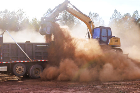 An excavator transfers large amount of dirt into truck, creating dust clouds in construction area under land removalの写真素材