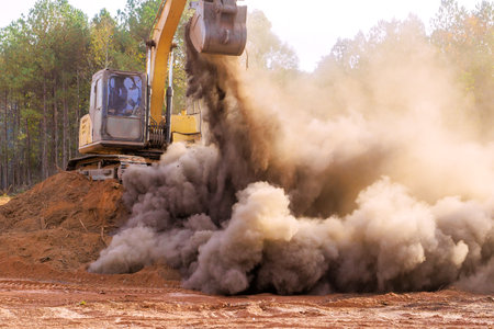 Large excavator is lifting load of dirt, generating significant amount of dust in clearing surrounded by construction work is ongoing at land preparationの写真素材
