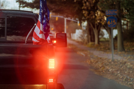 Pickup truck displaying an American flag travels along calm road surrounded by trees warm glow of dusk enhances peaceful atmosphere of autumn evening.の写真素材
