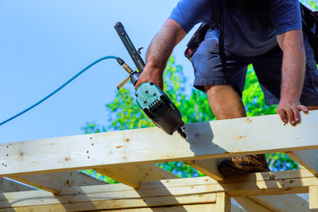 Construction worker fastens wooden beams while standing on roof, utilizing air power nail gun tool during construction at work areaの写真素材