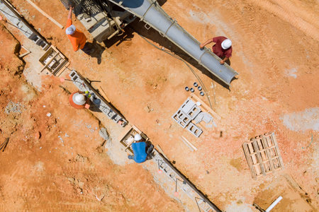 As concrete mixer truck pours concrete along foundational in spacious, reddish soil area, construction worker is observed using it.の写真素材