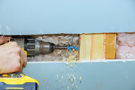 Worker operates power drill to securely fit insulation material between wooden studs in wall,...