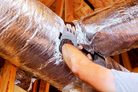 Contractor worker adjusts insulation around ductwork in residential attic, ensuring proper sealing efficiency.の写真素材