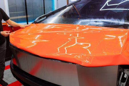 Worker applies bright orange vinyl wrap to car hood at customization workshop in car serviceの写真素材