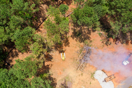 Heavy machinery clears trees in forested region while smoke rises from nearby debris, indicating active logging.の写真素材