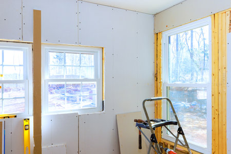 Construction work is underway with windows exposed gypsum drywall being installed in well lit space.