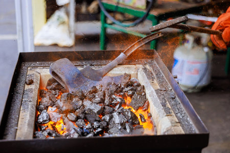 Hot coals glow as craftsman works with metal axe making, in busy forge, creating sparks in skilled...