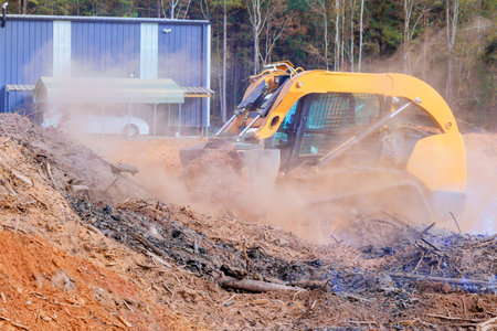 Heavy machinery removes debris, prepares land near commercial structure on dusty site surrounded by trees.の写真素材