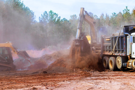 Heavy machinery works to excavate move dirt at construction site surrounded by trees on construction...