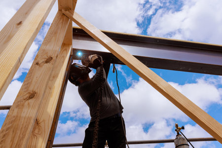 Craftsperson carefully welds metal beams while wearing protective gear under works day at job site.
