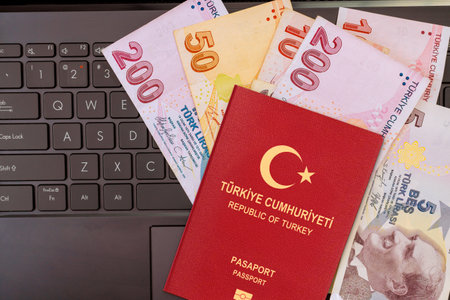 Turkish passport lies on laptop keyboard alongside various lira banknotes during travel preparation activity.の写真素材